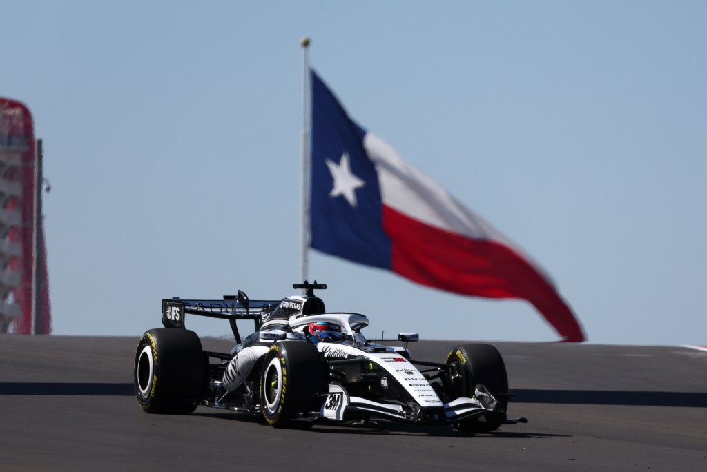 A Cadiallac Formula 1 racecar on a black track with a large Texas flag waving in the background against a blue sky.