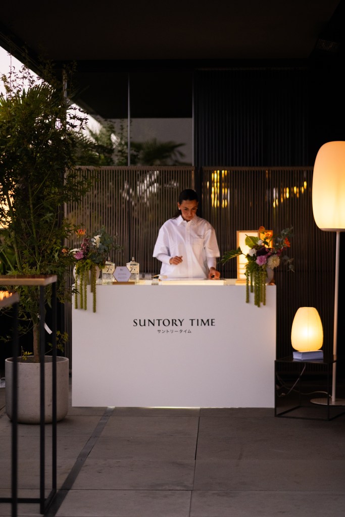 A woman wearing all white stands behind a white outdoor counter flanked by floral arrangements and "SUNTORY TIME" printed in English and Japanese on the facade.