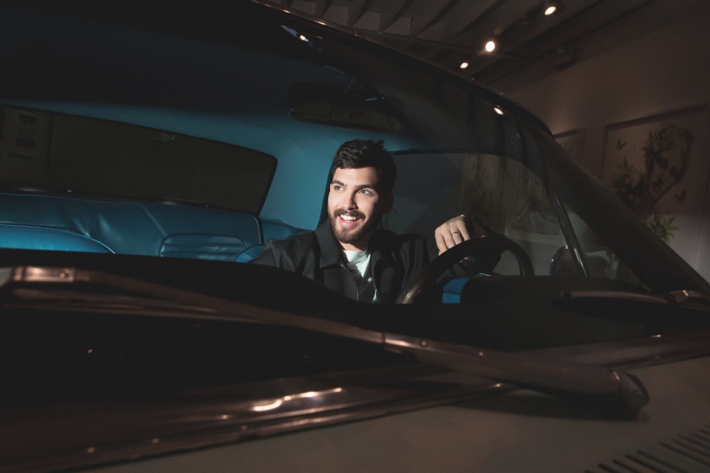 The artist KOZO sits in the drivers seat of a black car, smiling as he looks beyond the windshield.