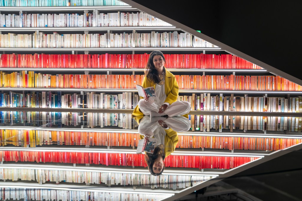 Artist Es Devlin sits cross legged on a mirrored floor wearing a yellow jacket and white pants with a rainbow of books on neat shelves behind her.