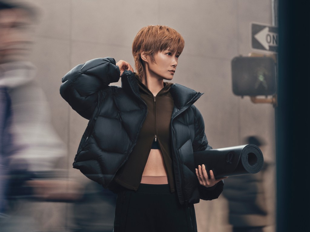 A woman wearing a cropped lululemon puffer with an arm raised touching her shoulder.
