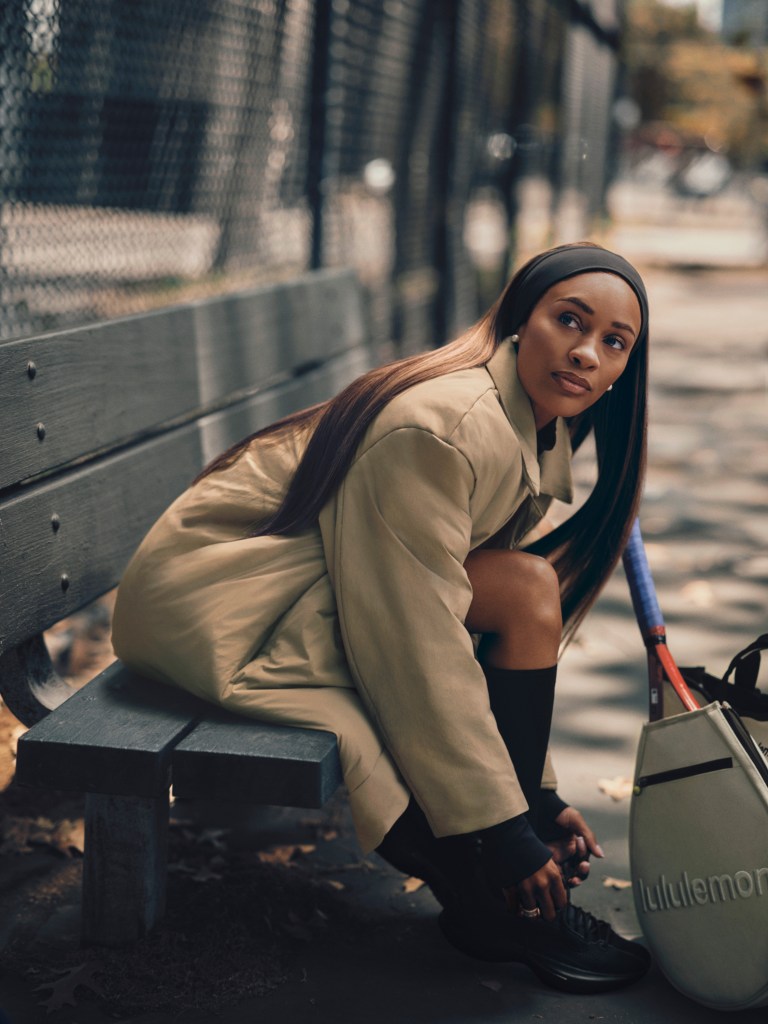 A woman wearing a long insulated lululemon jacket sits on a bench outside and leans over to tie her shoes.