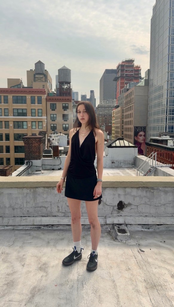 A woman wearing a black tank top, black mini skirt, white socks and a pair of black Nike sneakers stands on the rooftop of an apartment building with a city skyline behind her.