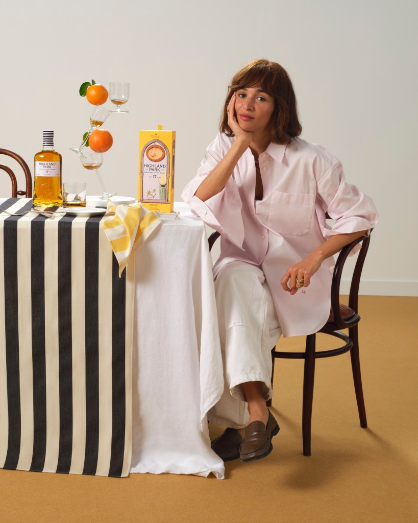 Laila Gohar sits cross legged at the end of a table with a bottle of Highland Park whisky and her sculpture made of glass and oranges.