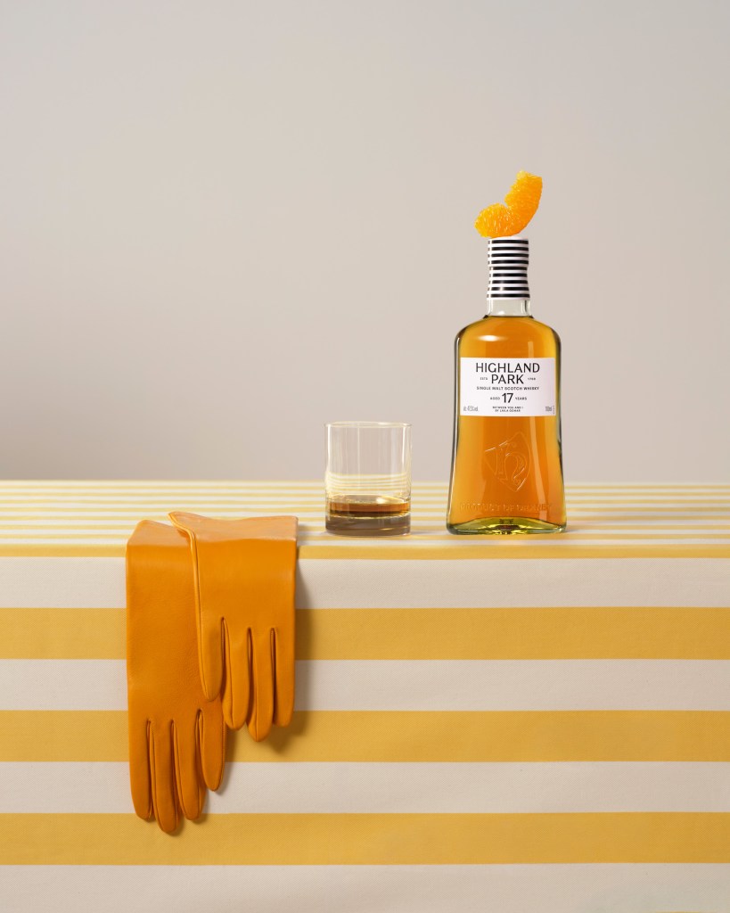 A bottle of Highland Park whisky with an orange slice on top with a glass and a yellow pair of gloves next to it.