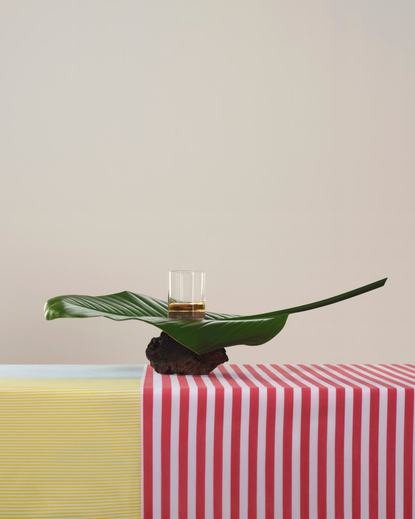 A table with two different patterned tablecloths and a sculptural leaf with a glass of whisky on it.