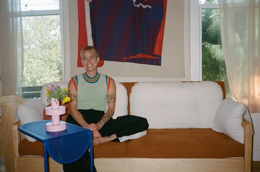 A person with a buzzed head and light green shirt sits casually on a cream couch flanked by windows, with a large red and navy tapestry hanging behind them.