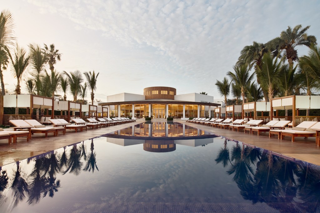 A large outdoor swimming pool looks onto a grand hotel facade flanked by trees.