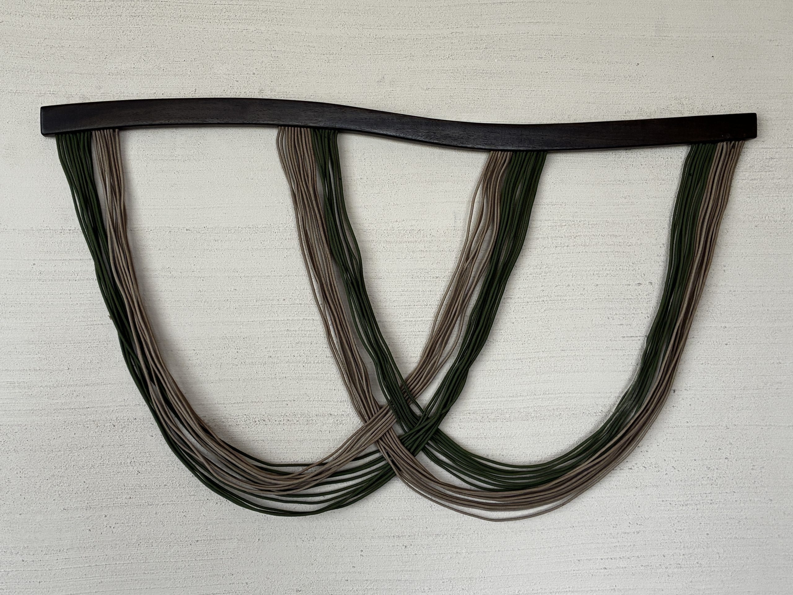 A textile artwork of two groups of yarns hanging from a stick, intersecting in the middle, on a white textured wall.