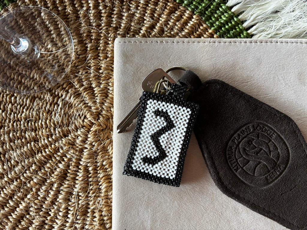 A overhead shot of a black and white beaded keychain that says "3" sitting on top of a leather booklet and a found woven placemat.
