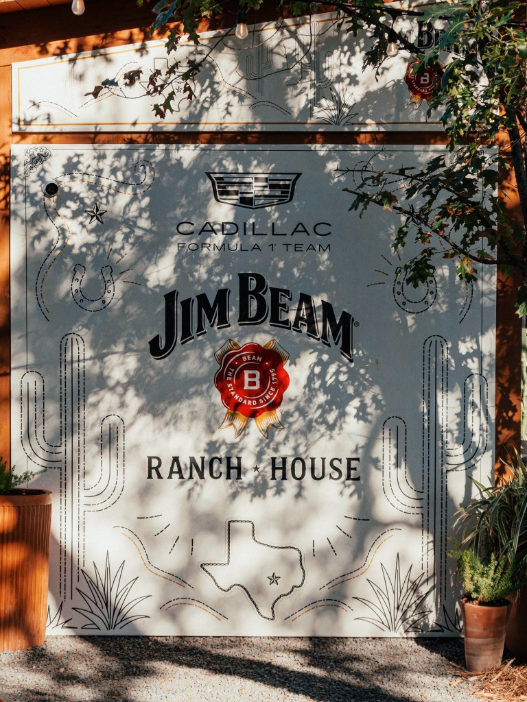 A large white sign that says "Cadillac Formula 1 Team, Jim Beam Ranch House" with illustrations of horseshoes, cacti and an outline of the state of Texas.