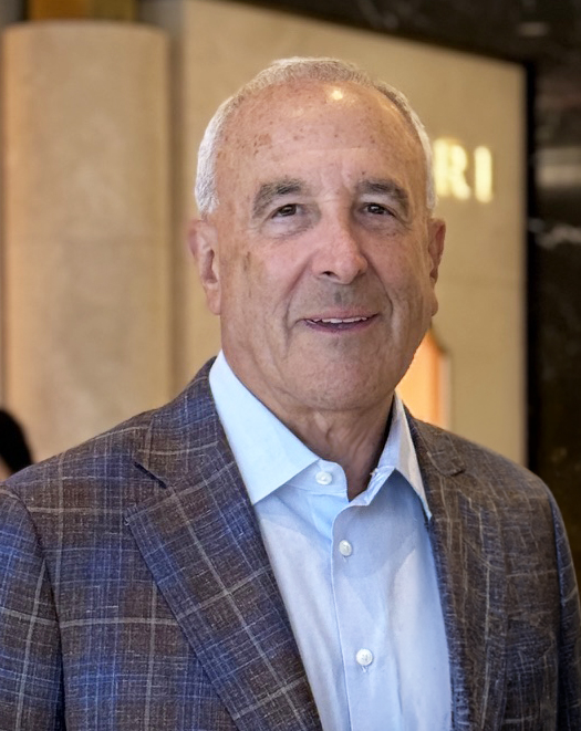 A tight crop of a man wearing a plaid blazer and light blue button-up shirt with a slight smile.