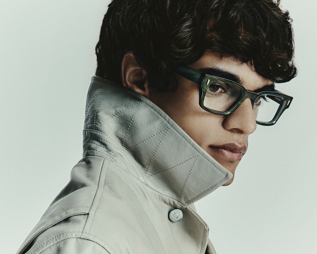 A model wearing a chunky black pair of Mortgenthal Frederics eyeglasses and a light-colored jacket with an upturned collar.