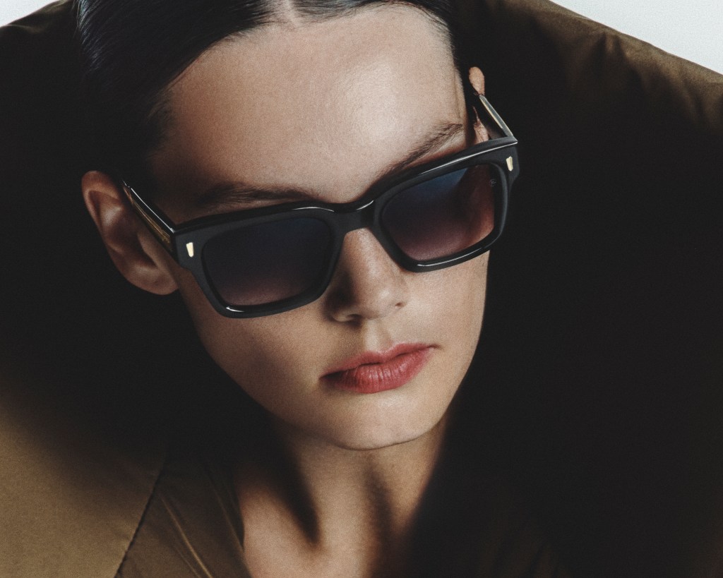 A model with red lips and slicked back hair wear a large pair of sunglasses.