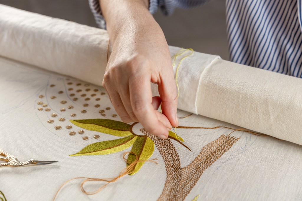 A close up of a woman's hand holding a needle and thread on top of a large embroidered piece of cream fabric. 