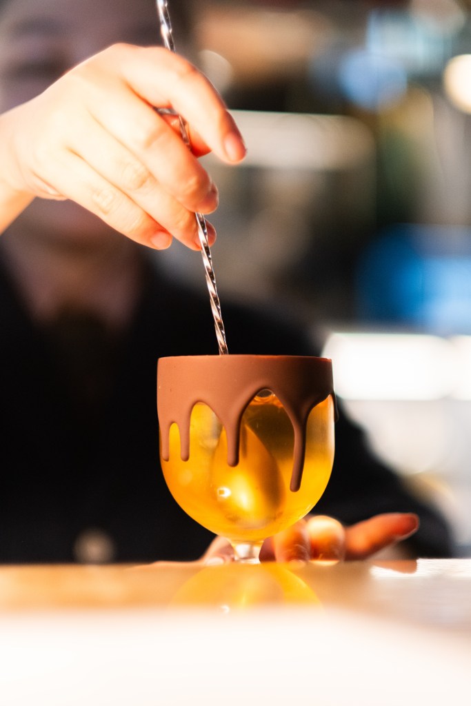 A hand stirring a chocolate-rimmed cocktail in a golden goblet.