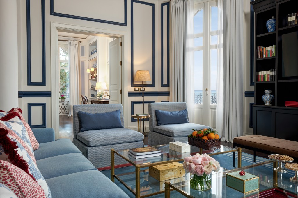A living room with blue chairs, white walls with navy trim, two glass and brass tables with flowers, fruit and treats sitting on them, and a tall window overlooking the sea outside.