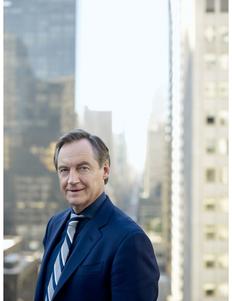 A man wearing a striped tie and a dark navy suit stands in front of a cityscape of tall office buildings behind him.