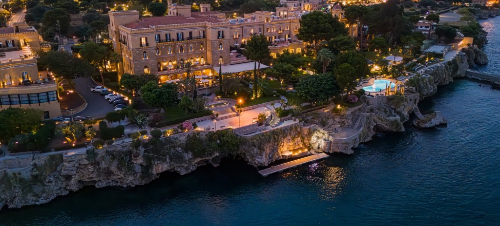 An aerial nighttime shot of Villa Igiea on the coast with warm lights illuminating the property.