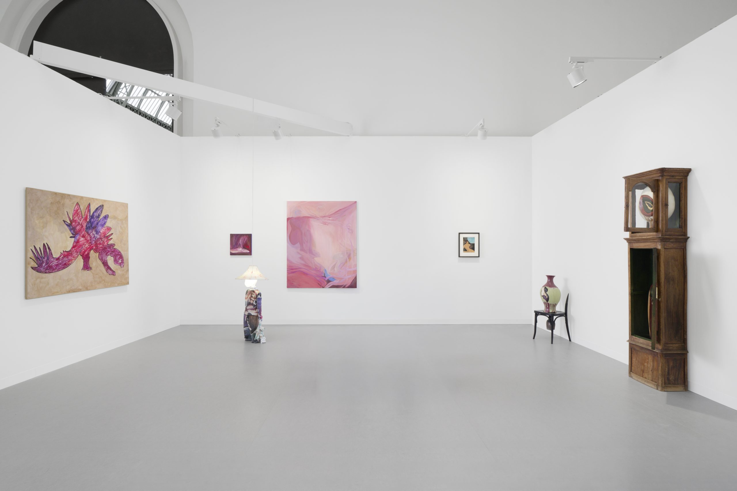 A three-walled gallery room with a gray floor and multiple artworks displayed, including a child-like sculpture with an illuminated head.