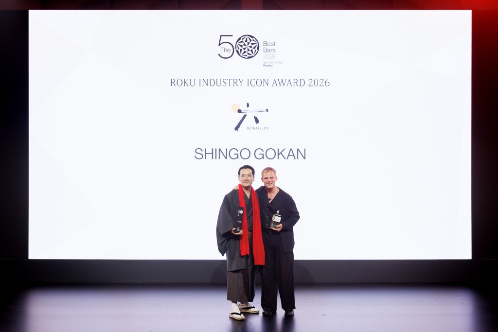 Two people standing on the stage holding an award and a bottle of gin with "ROKU INDUSTRY ICON AWARD 2026: Shingo Gokan" projected behind them on a white screen.