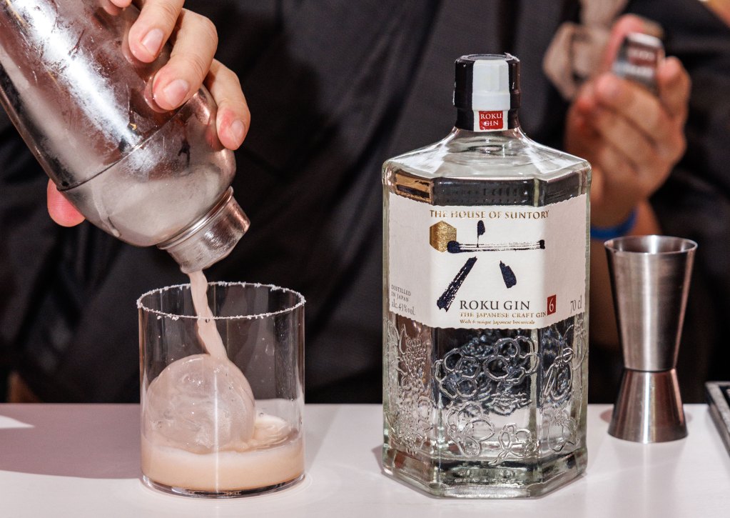 A hand pouring a cocktail into a rocks glass with a round ice cube next to a bottle of Roku Gin.