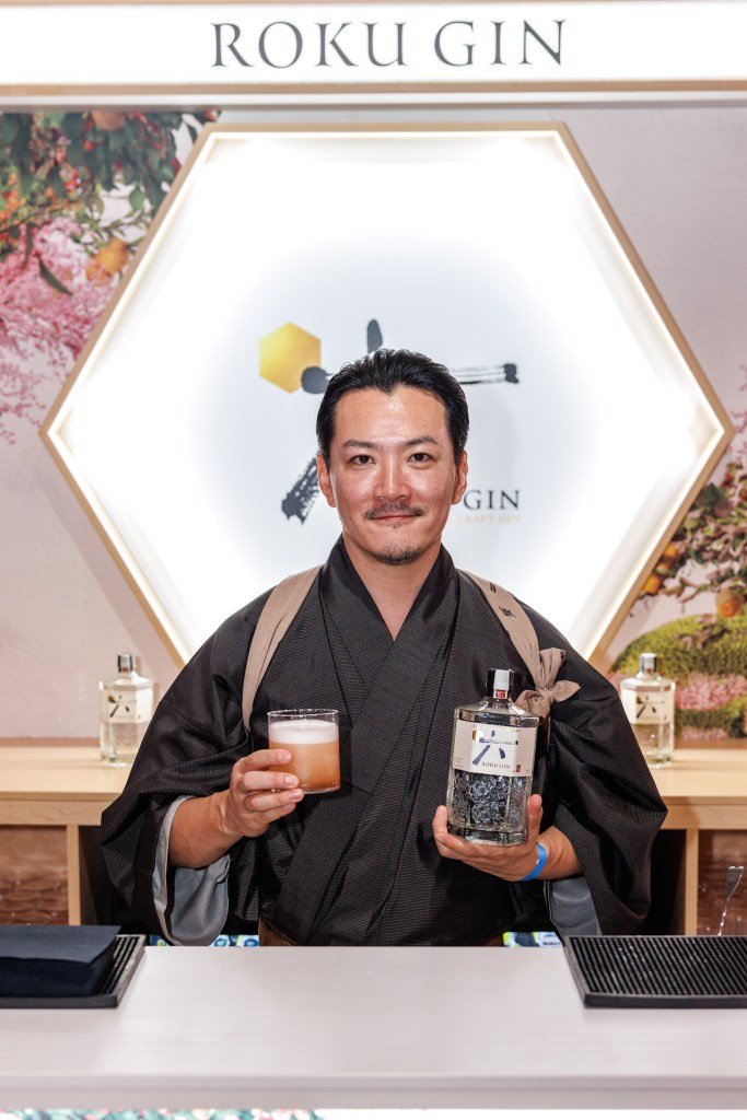 Shingo Gokan standing in front of a hexagonal sight that says Roku Gin as he holds a cocktail and a gin bottle.