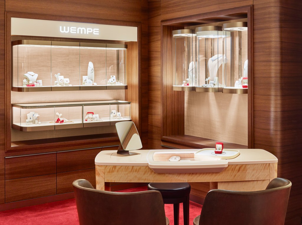 A table with two chairs in front of it and two tall jewelry cases behind it in a room with soft light and red carpet.