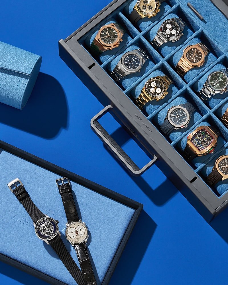 A gunmetal gray watch case full of watches sits on a bright blue surface with two other watches on a light blue tray. 