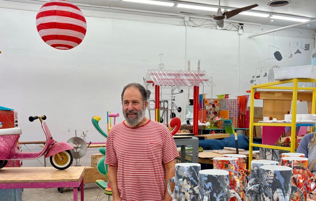 A bearded man wearing a red-and-white striped tee shirt smiles in front of his studio that has many whimsical and brightly colored ceramic sculptures on display.